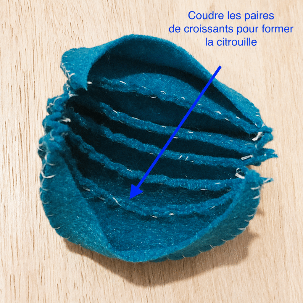 Étape de couture d’une citrouille en feutrine bleue, avec plusieurs croissants cousus ensemble sur une table en bois clair.